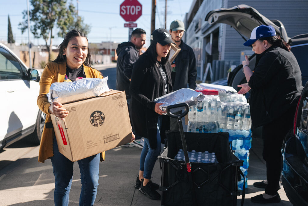 A group of people is engaged in a community effort, carrying boxes and supplies. One person holds a Starbucks box, while others manage bags and bottles of water. A cart filled with water bottles is nearby, and a stop sign is visible in the background.