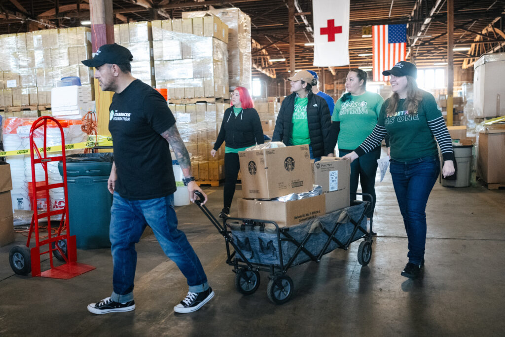 A group of volunteers is moving boxes on a cart in a warehouse. They wear matching green shirts and are engaged in a community service activity. The background features stacked boxes and flags.