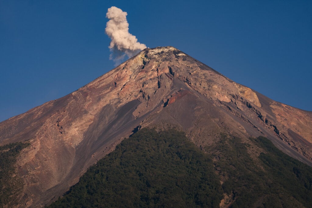 A volcano with a plume of smoke rising from its summit, surrounded by rocky terrain and lush greenery at its base. The sky is clear and blue.
