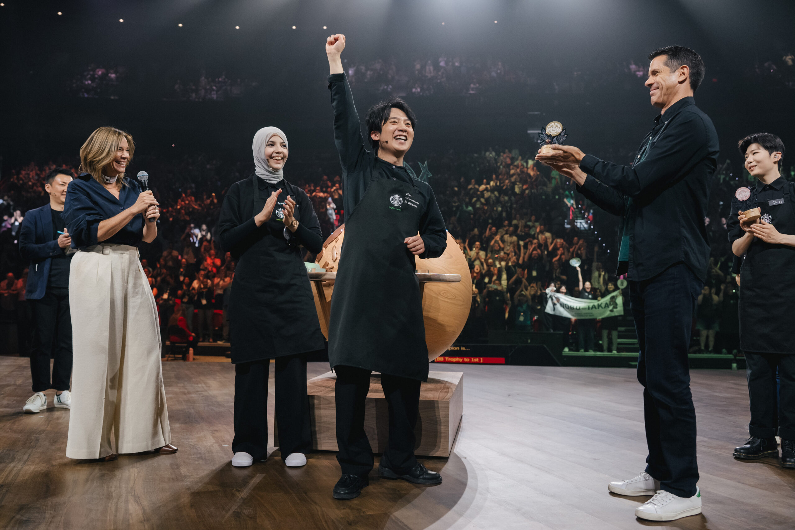 A celebration scene with three individuals on stage. One person, wearing a black apron, raises a fist in triumph, while another claps. A trophy is being presented by a third person. The background features a cheering crowd.