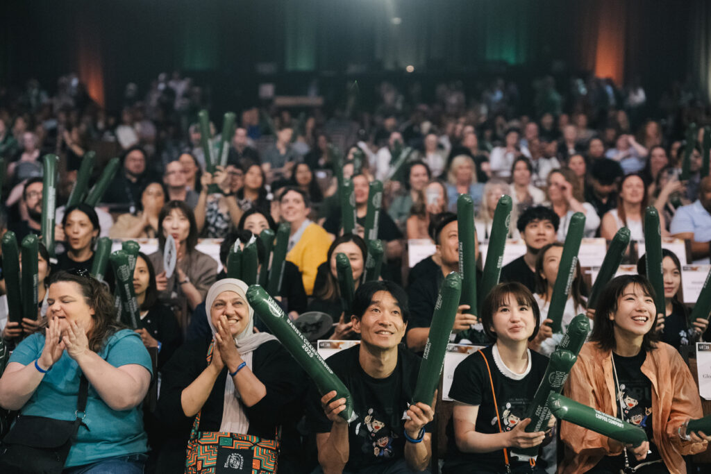 A lively audience is cheering, holding green inflatable sticks. People are smiling and clapping, creating an energetic atmosphere.