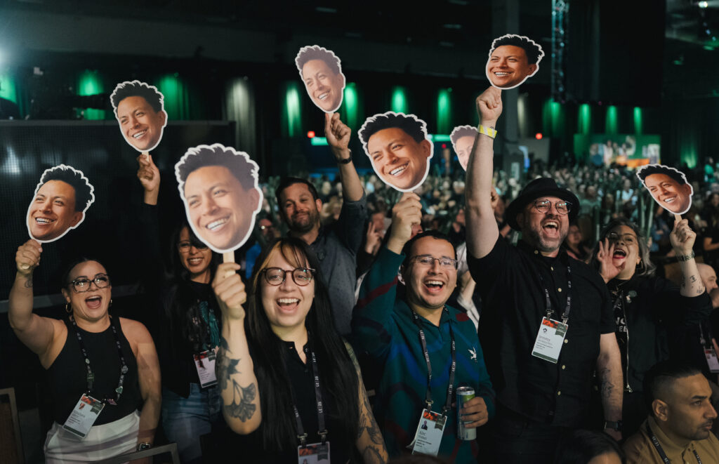 A lively crowd holds up large cutouts of a smiling face, showing excitement and enthusiasm. People are smiling and cheering, with some wearing name badges.