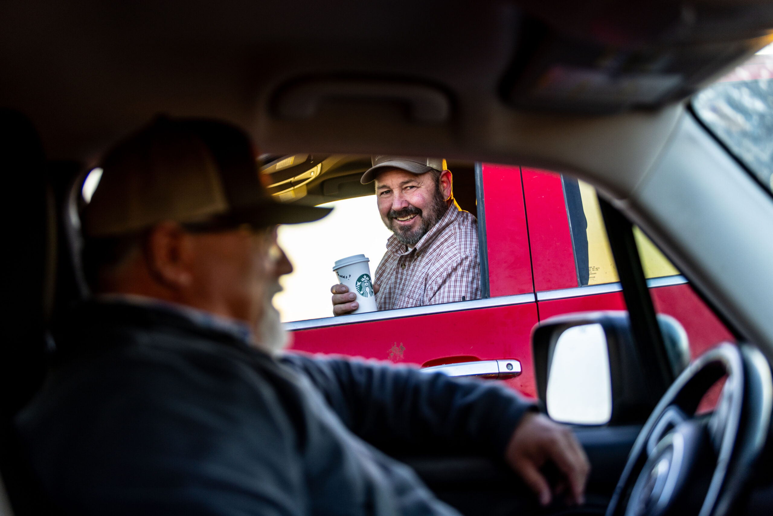 A man with a beard and a plaid shirt smiles from the driver's side of a red vehicle, holding a Starbucks cup. Another person is partially visible in the foreground.