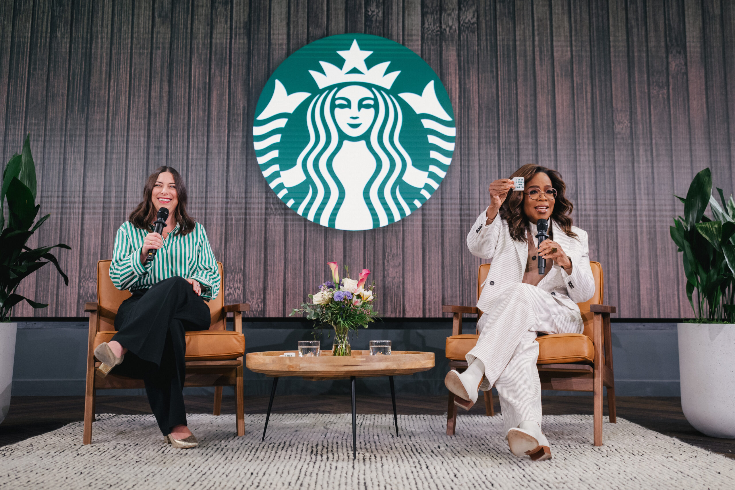Two women are seated on stage, one holding a microphone and displaying a card, while the other smiles and holds a microphone. Behind them is a large Starbucks logo on a wooden backdrop. A small floral arrangement and glasses of water are on a round wooden table between them.