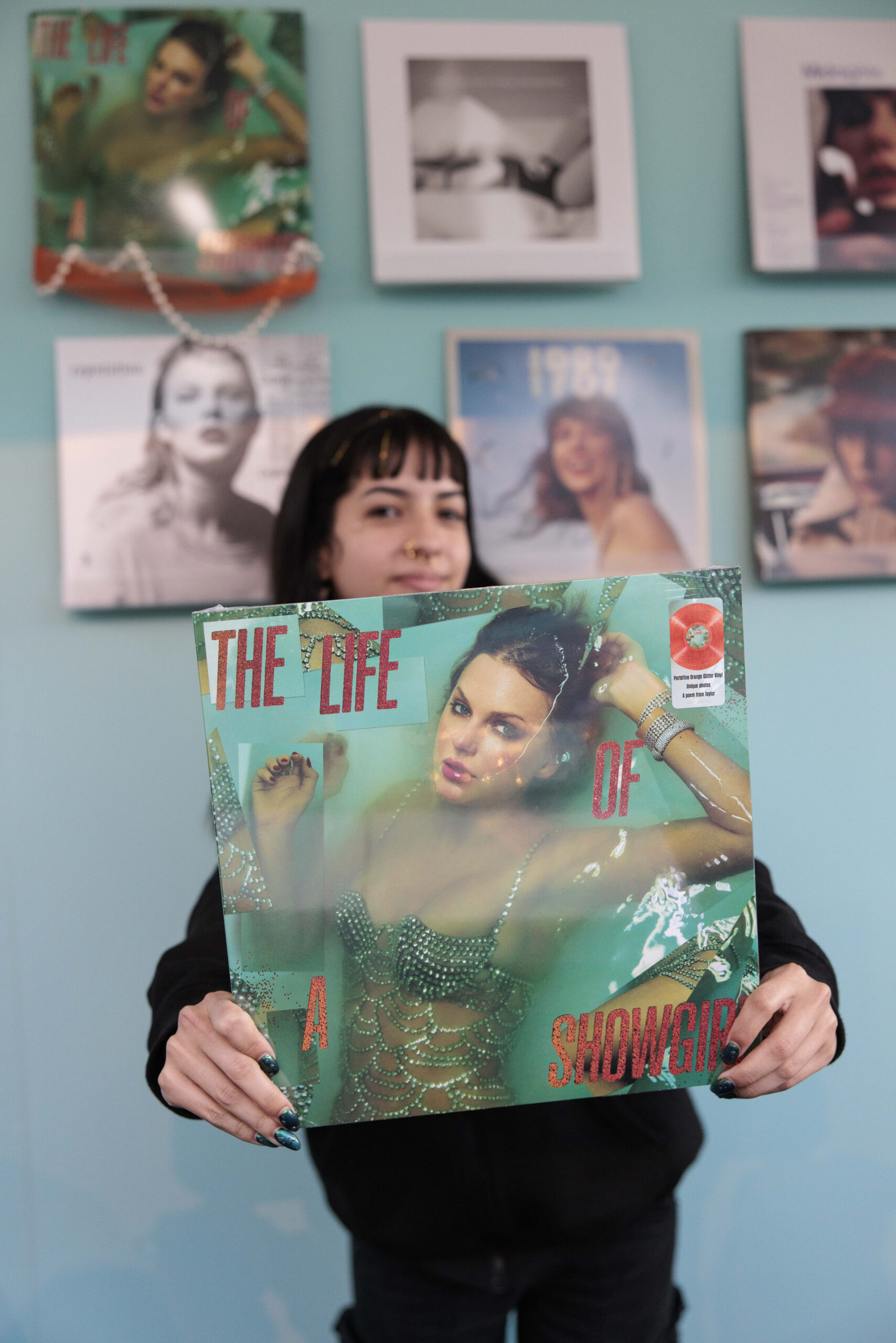A person holds a vinyl record featuring a glamorous image of a woman in a jeweled outfit, with the title "THE LIFE OF A SHOWGIRL." The background includes framed album covers on a light blue wall.