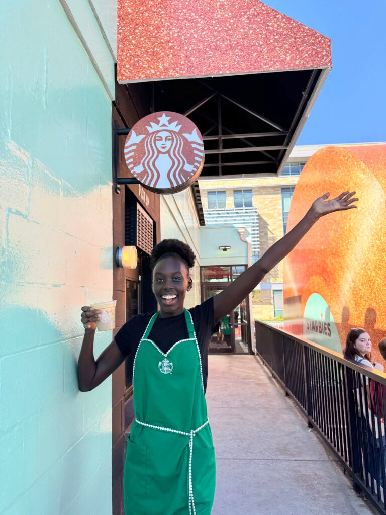 A person in a green Starbucks apron is smiling and waving, holding a drink. The background features a colorful wall and a Starbucks sign.