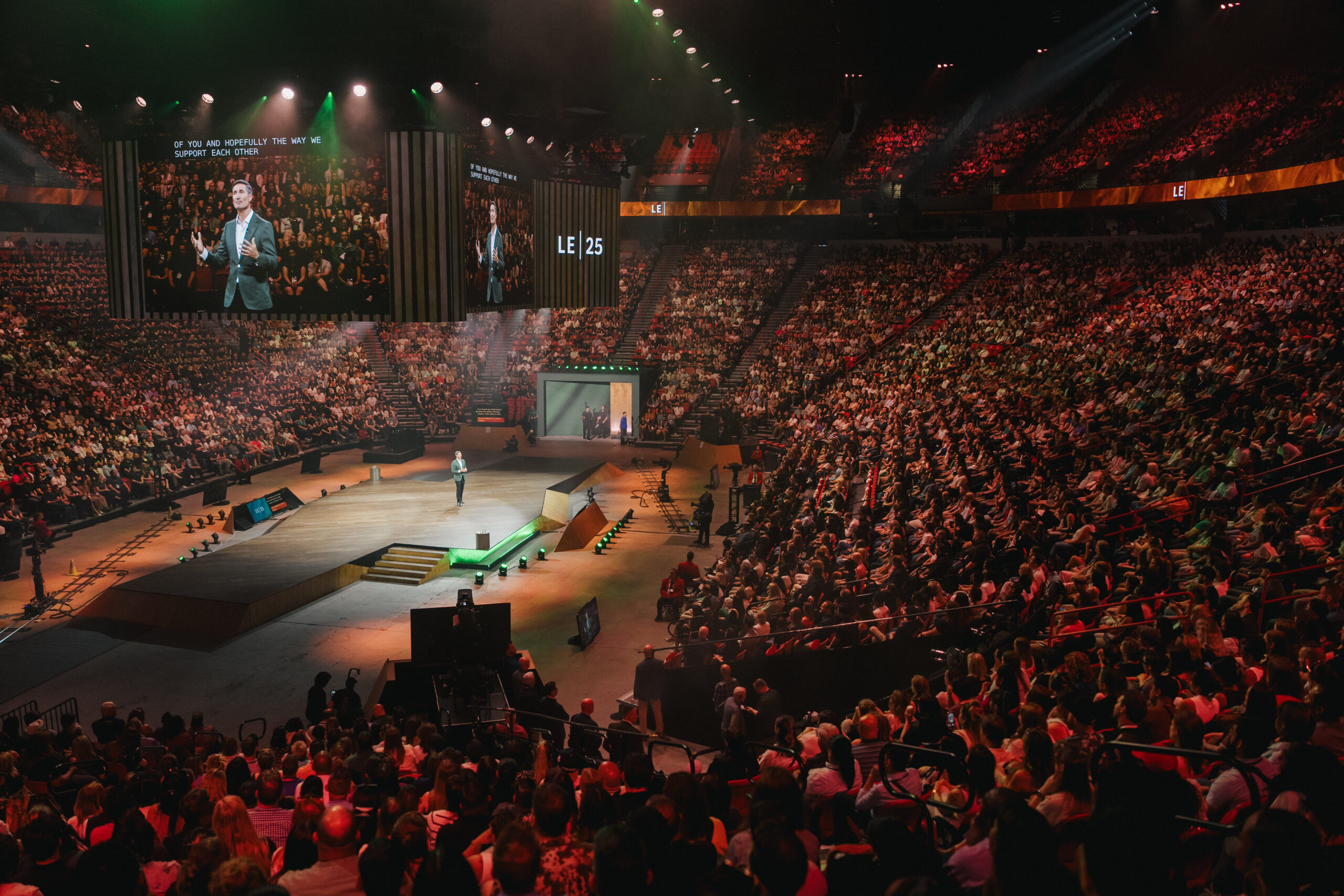 A large audience is gathered in an arena for a conference. A speaker stands on stage, with multiple screens displaying visuals. The crowd is engaged, and the venue is brightly lit with green accents.