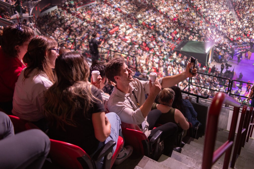 A group of people is seated in a crowded arena, with one man taking a selfie while holding a drink. The audience is engaged, with many visible in the background.