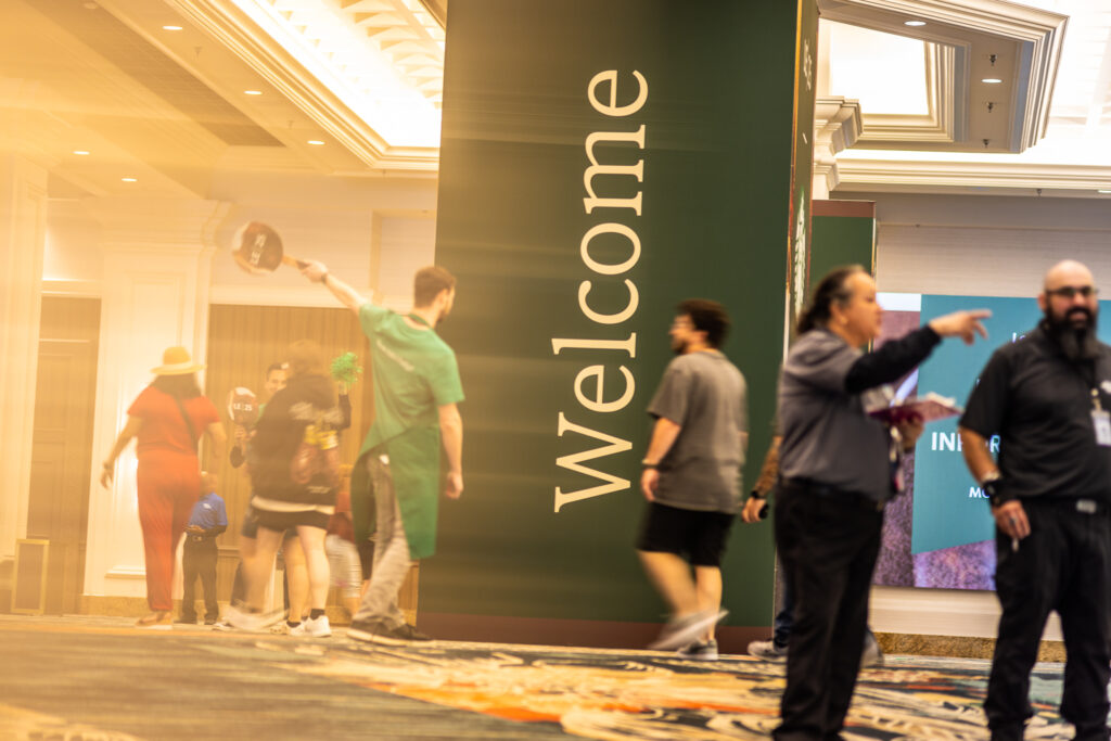 A large "Welcome" sign is prominently displayed in a busy hallway. People are walking by, some in casual attire, while a staff member in a green shirt waves a sign. The atmosphere is lively and welcoming.