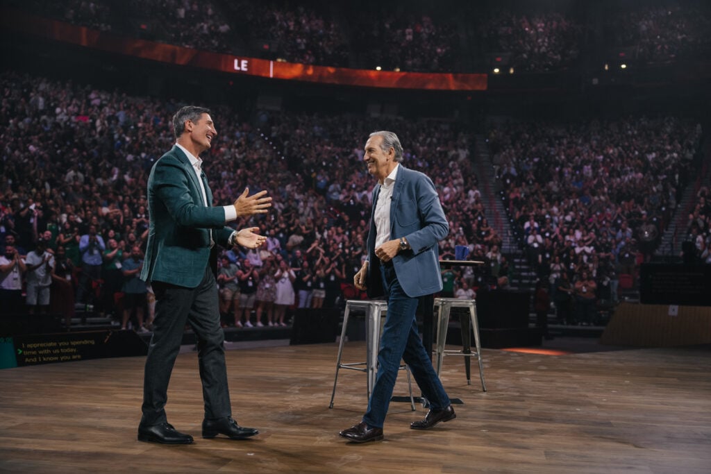 Two men are on stage, engaging with each other and smiling. A large audience is visible in the background, with many people applauding. There are two stools on stage, and the setting appears to be a lively event or conference.