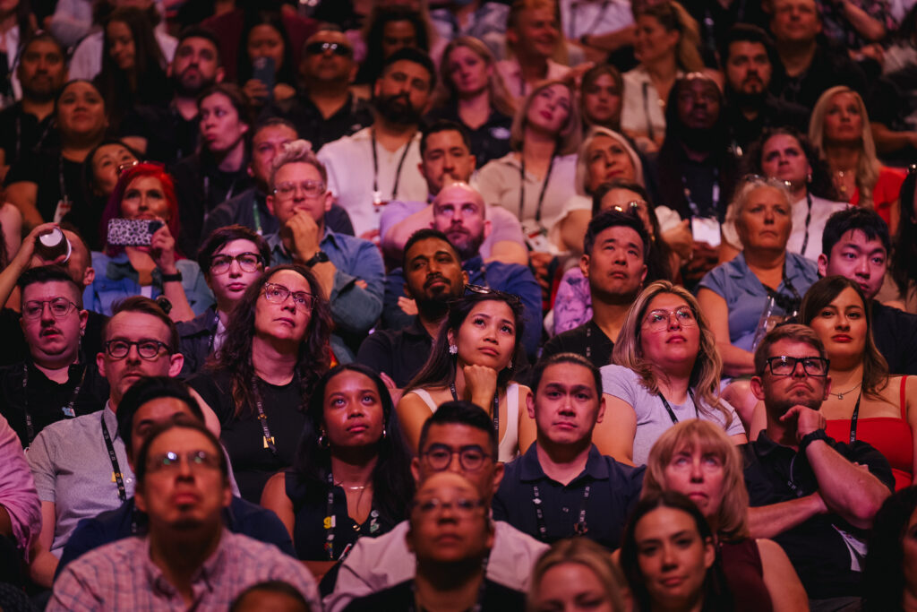 A diverse audience is seated closely together, looking up with expressions of curiosity and contemplation. Many are wearing lanyards, indicating they are likely attending an event or conference. The lighting creates a warm atmosphere, highlighting their engaged faces.