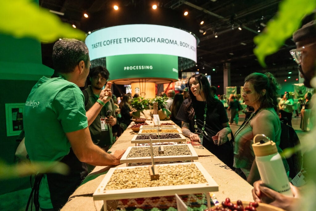 A coffee tasting event with a presenter discussing various coffee beans displayed on a table. Attendees engage and observe.