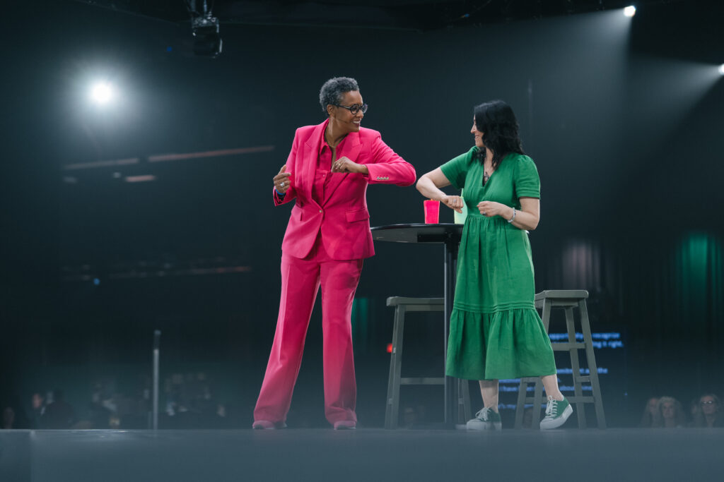 Two women are on stage, one in a bright pink suit and the other in a green dress. They are interacting playfully, with a small table and a red cup between them. The background is dimly lit, focusing on their lively engagement.