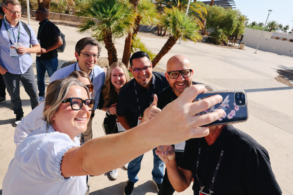 A group of six people is taking a selfie outdoors, smiling and posing with thumbs up. Palm trees and a sunny background are visible.