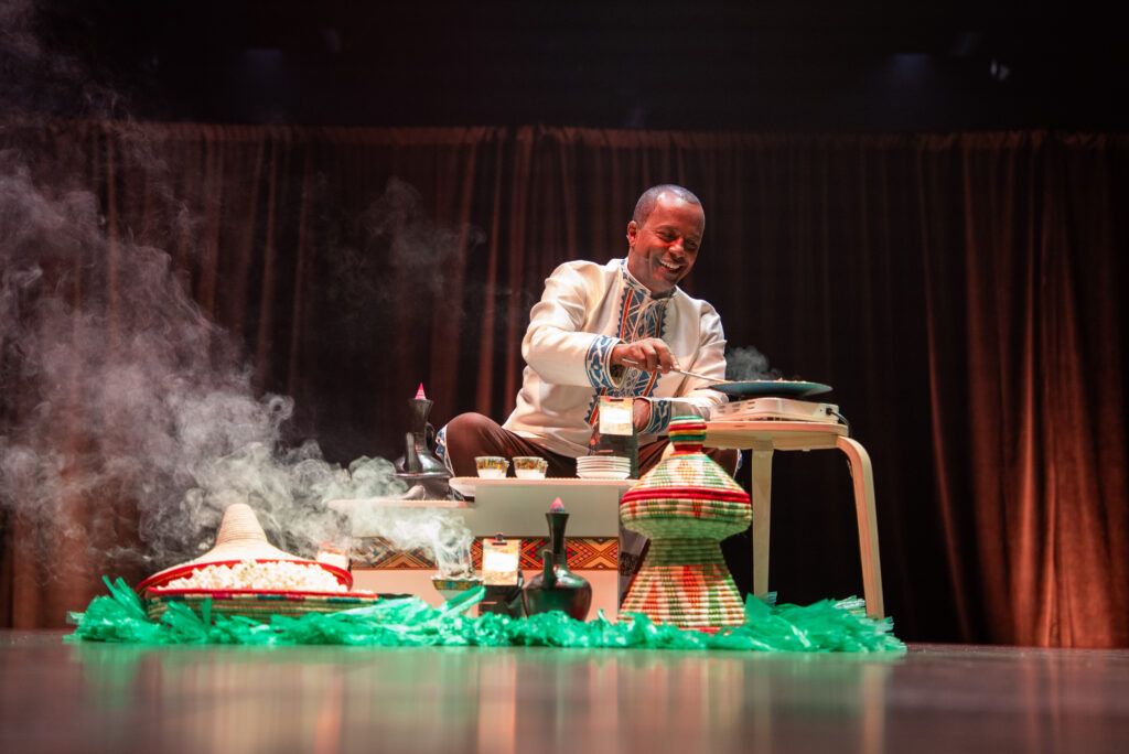 A man in traditional attire prepares food on a small table, surrounded by colorful dishes and decorative items. Smoke rises from the cooking area, creating an inviting atmosphere.