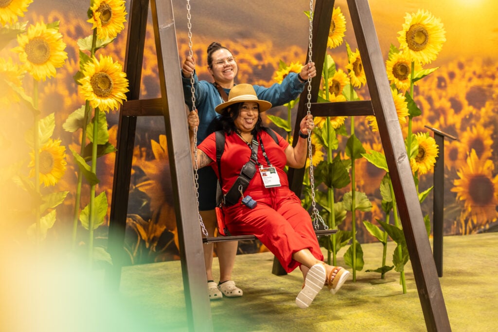 Two women are enjoying a swing set surrounded by large sunflowers. One woman is seated on the swing, smiling, while the other stands behind her, playfully pushing the swing. The background features a vibrant sunflower field.