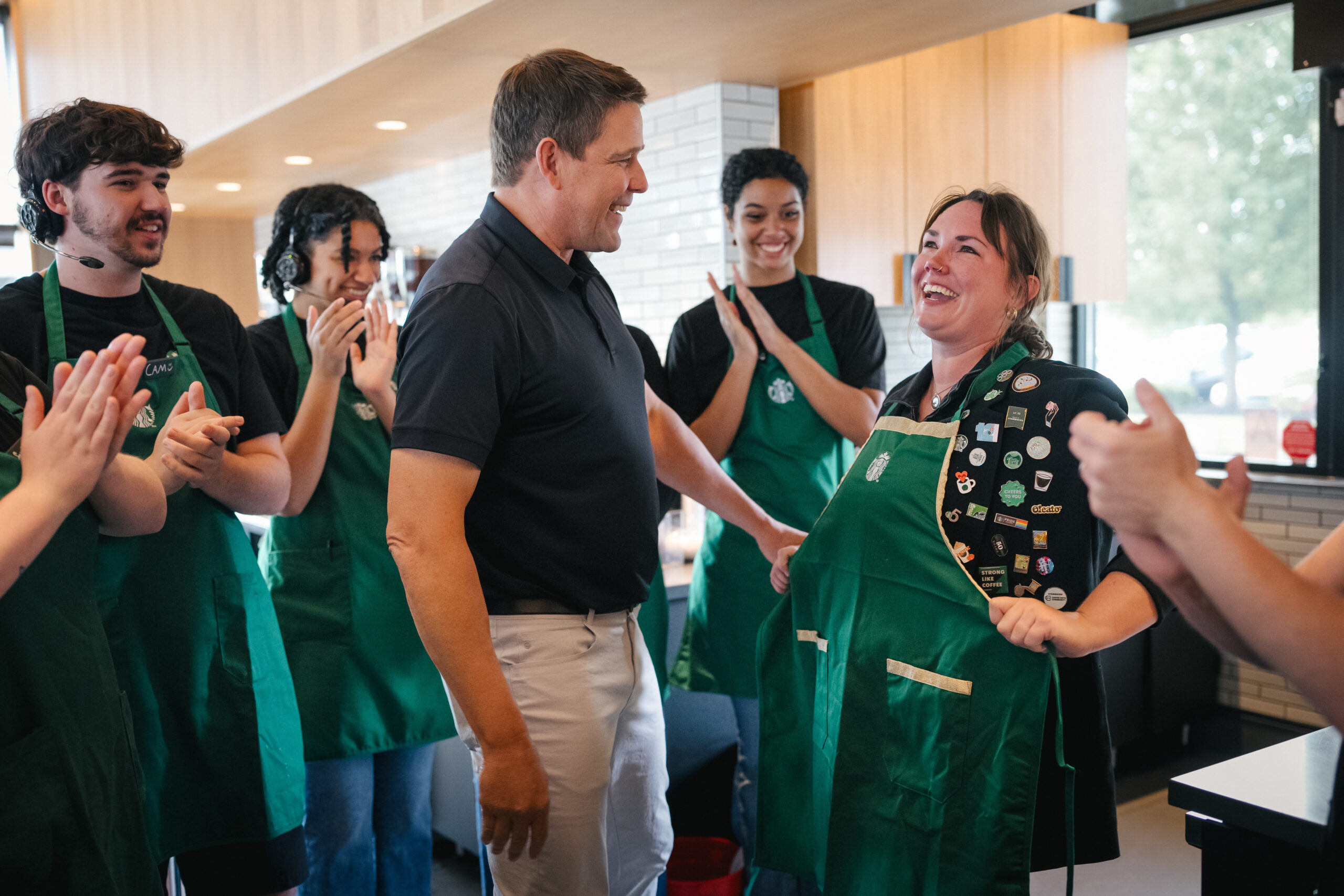 A group of baristas in green aprons are celebrating with a smiling woman wearing a decorated apron. A man in a black shirt engages with her, while others applaud joyfully in a coffee shop setting.