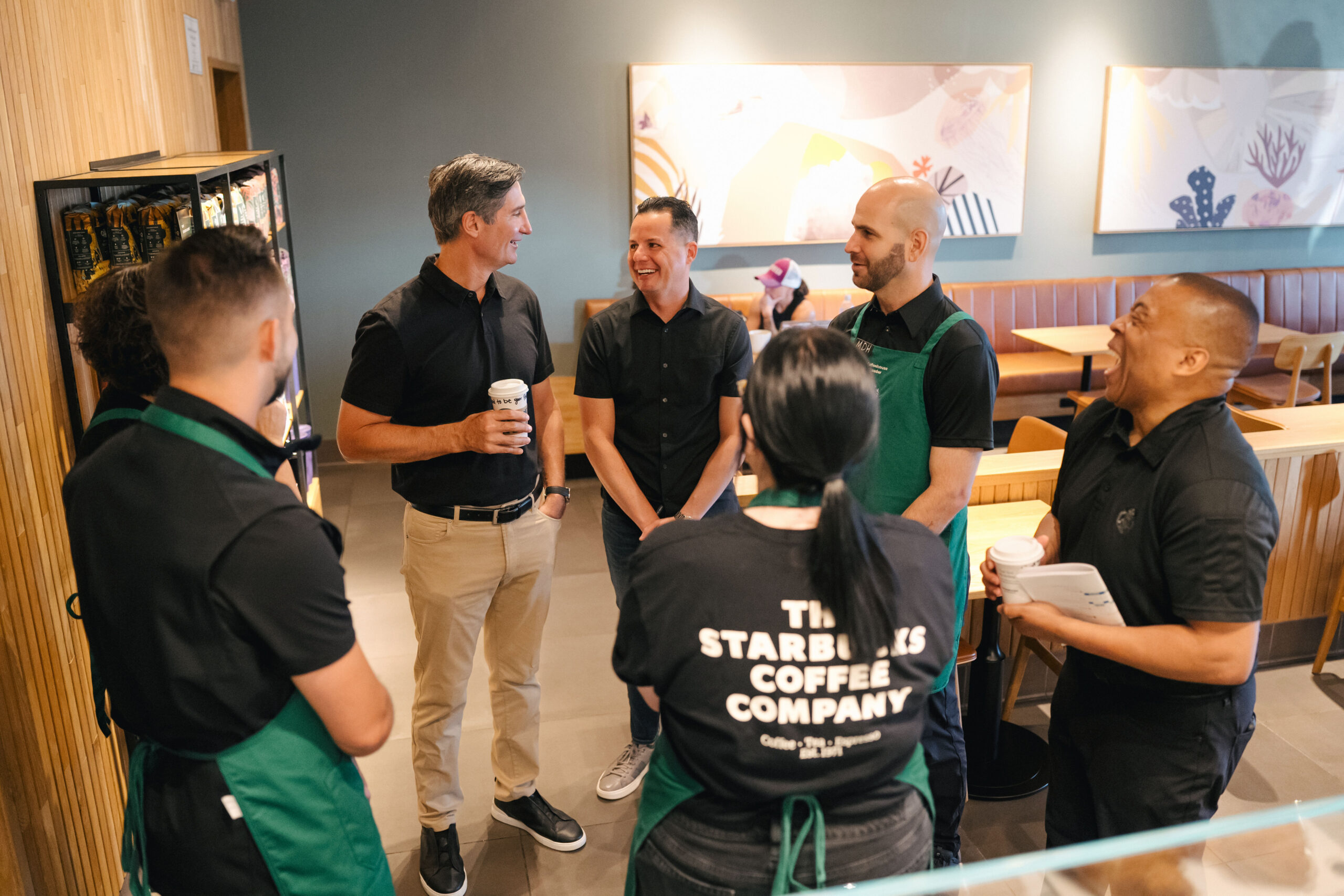 A group of six individuals, dressed in black shirts and green aprons, engage in conversation in a coffee shop. One person holds a coffee cup, while others listen attentively. The setting features modern decor with seating areas in the background.