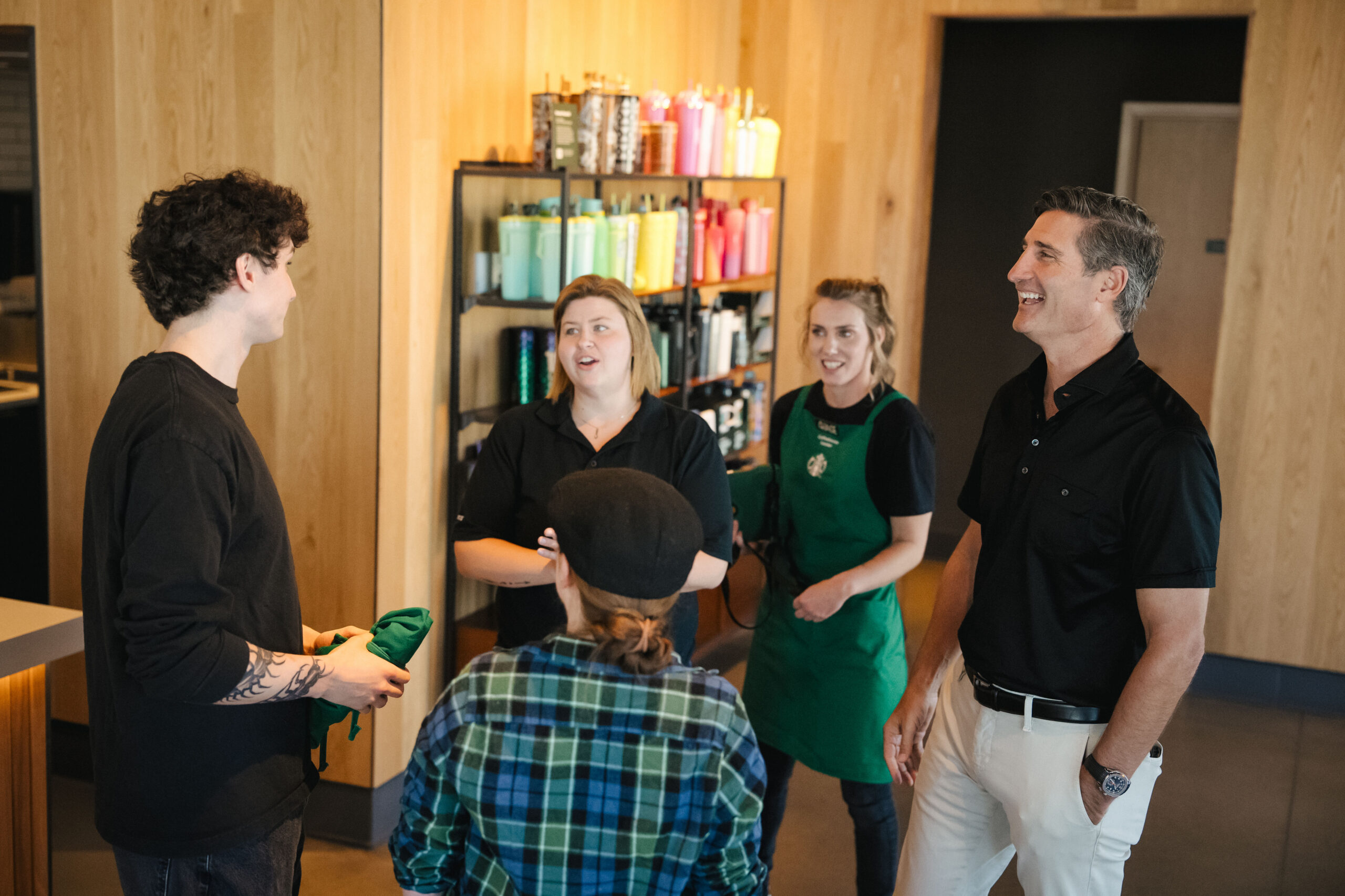 A group of four people is engaged in conversation inside a coffee shop. Two are wearing black shirts, one in a green apron, and another in a plaid shirt. A shelf in the background displays colorful drinkware.