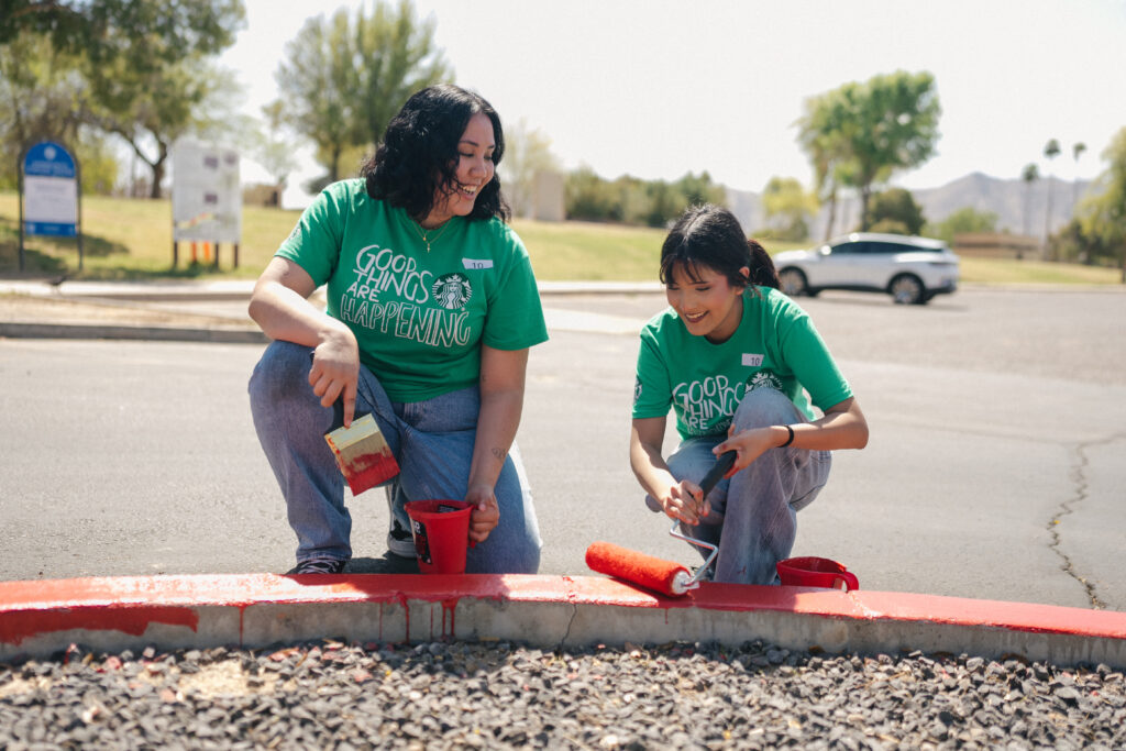 Two individuals in green shirts are painting a red curb. One is using a paint roller, while the other holds a paintbrush and bucket.