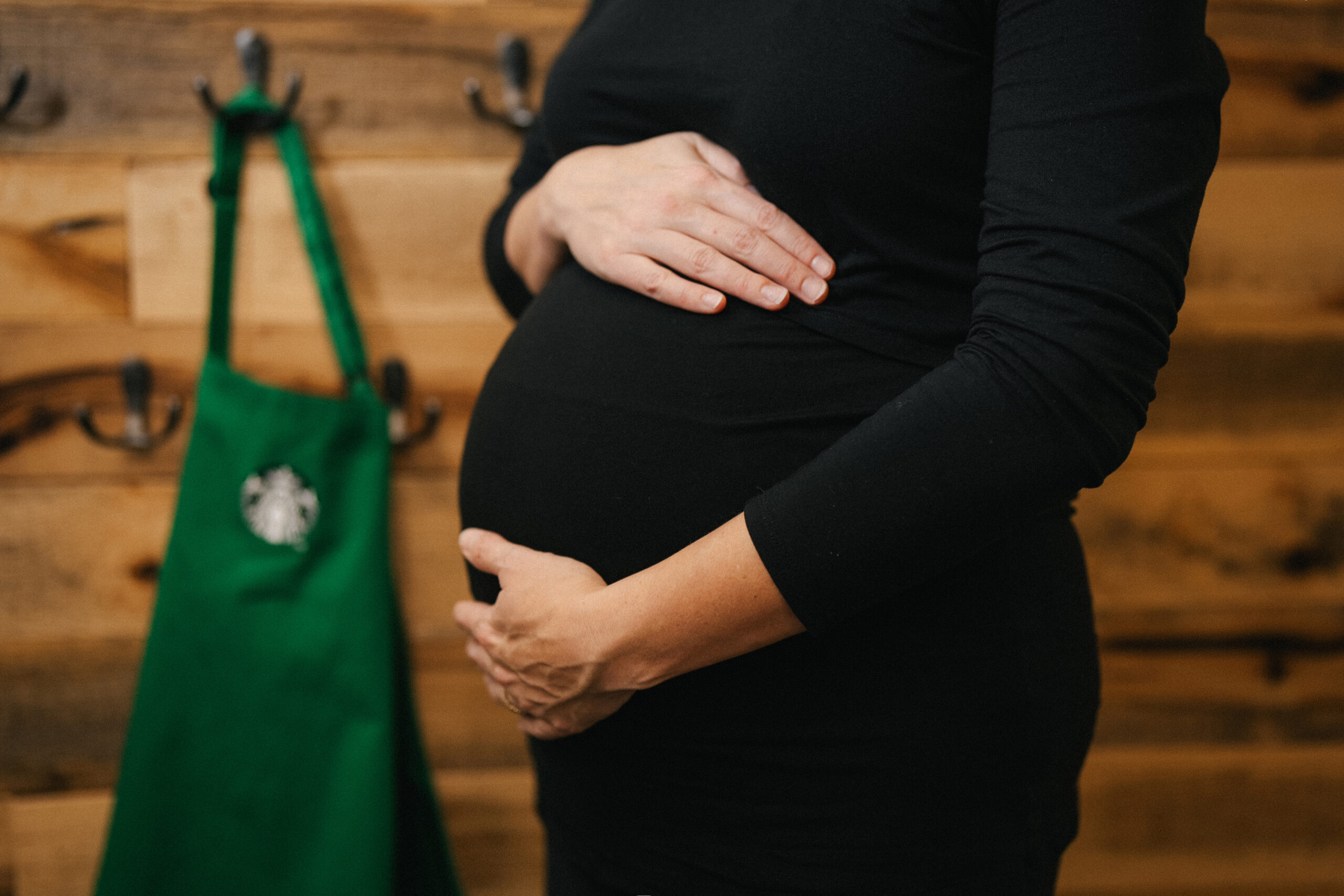 A close-up of a pregnant person's belly, with one hand resting on top and the other cradling the bottom. A green bag is visible in the background.