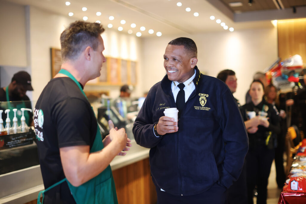 A police chief in a navy fleece jacket smiles while holding a coffee cup, engaging in conversation with a barista in a café.