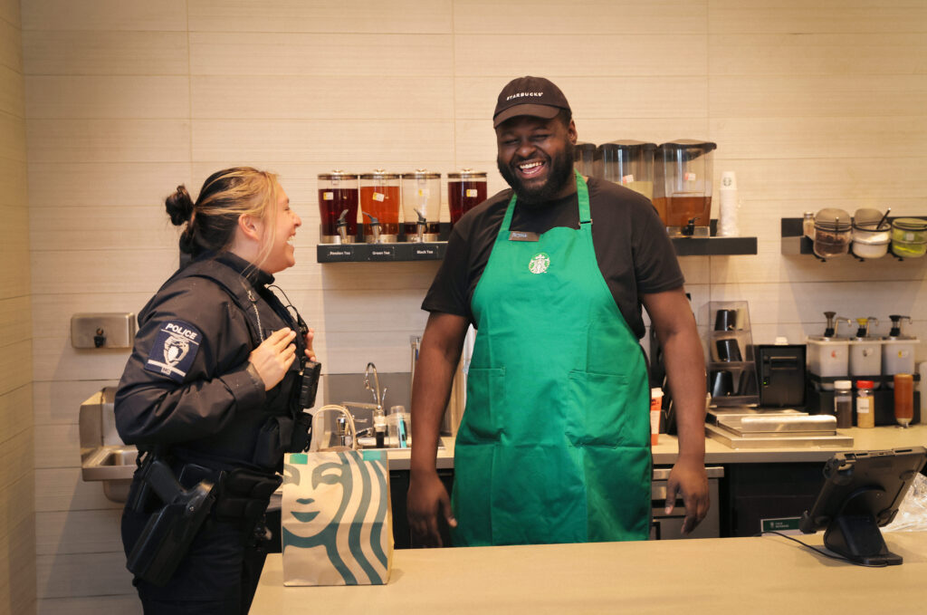 A police officer and a barista are laughing together at a Starbucks counter. The barista wears a green apron, and there's a Starbucks bag on the counter. Behind them, various tea dispensers are visible.