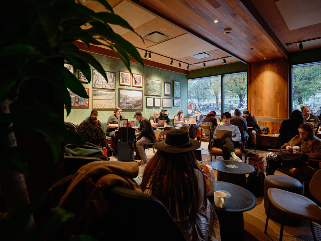 A cozy café scene with patrons seated at tables, some working on laptops. The walls are adorned with framed artwork, and large windows provide natural light. A few people are engaged in conversation, while others enjoy their drinks.