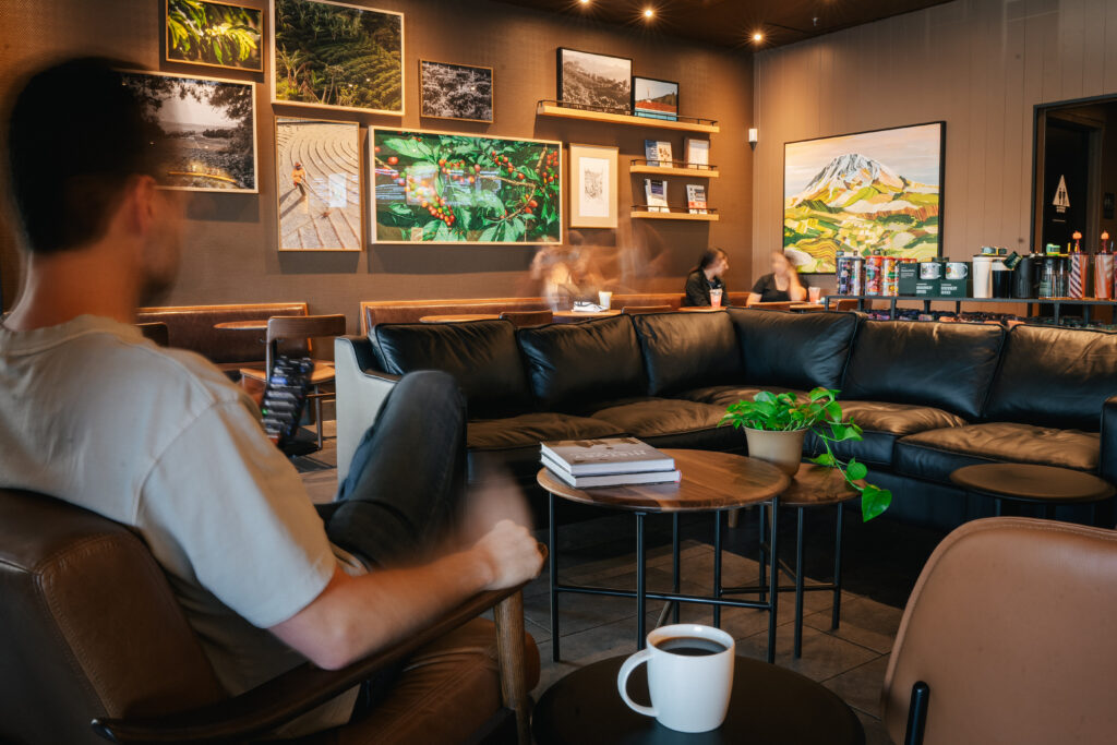 A cozy café interior features a black leather sectional sofa, wooden tables, and a potted plant. Artwork adorns the walls, and patrons are seated, enjoying drinks. A coffee cup sits on a table in the foreground.