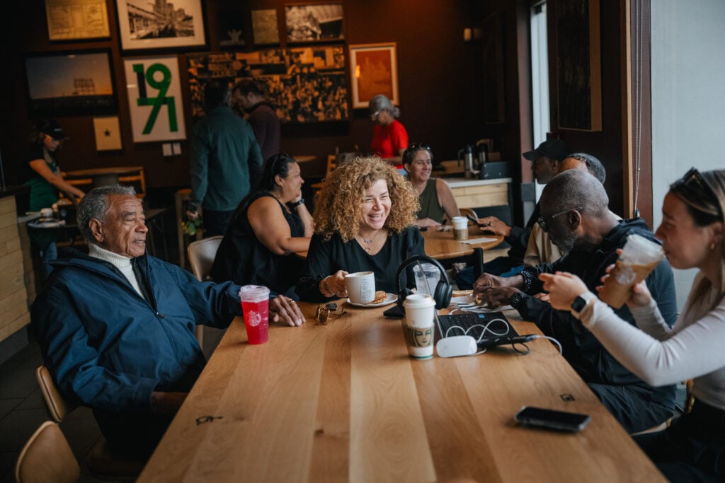 A lively café scene with several people seated around a wooden table. A woman with curly hair is smiling and holding a cup, while others engage in conversation. Drinks and snacks are visible on the table, along with electronic devices. The atmosphere appears warm and social.