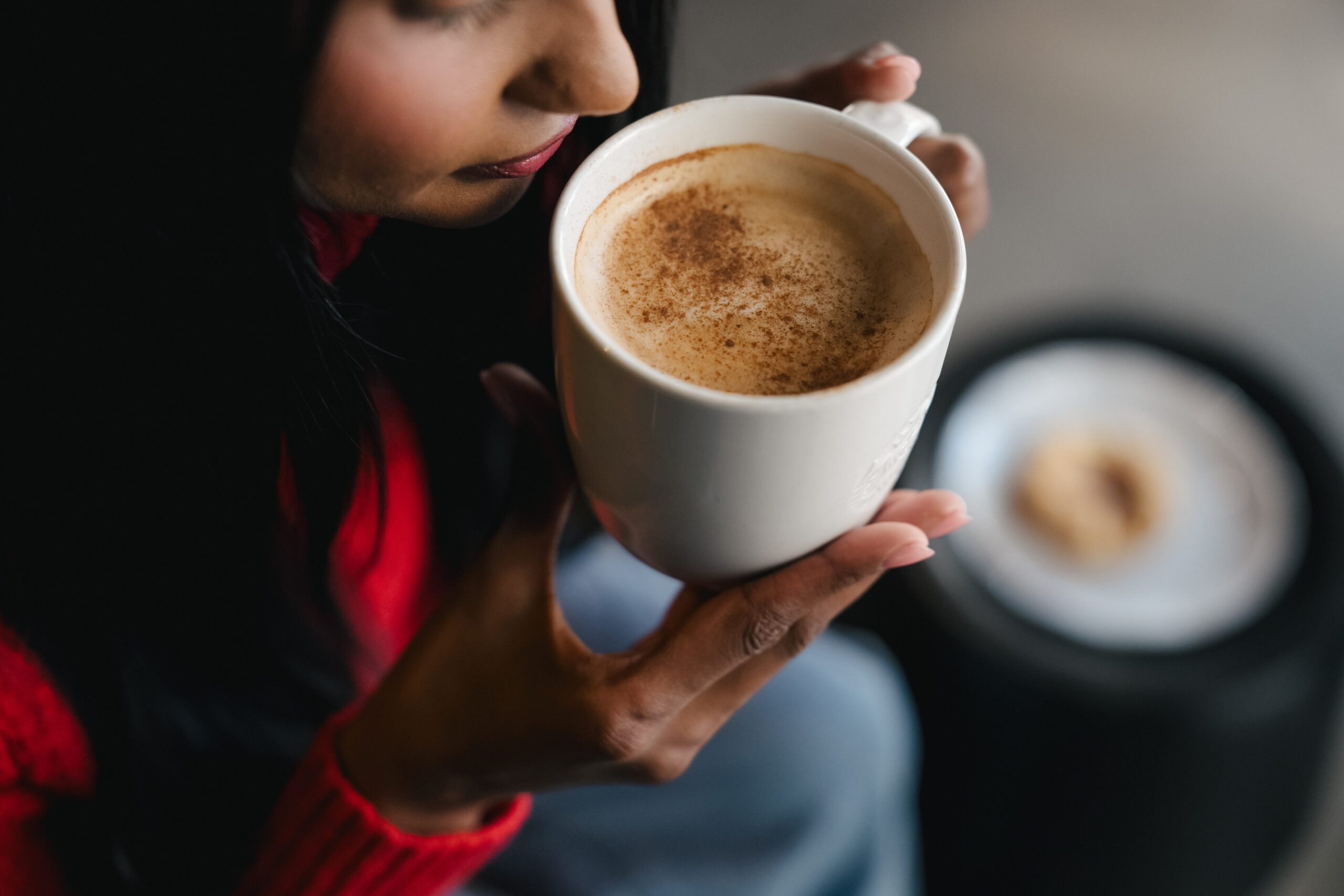 A person holds a white mug filled with a frothy beverage topped with a sprinkle of cinnamon. A plate with a cookie is visible in the background.