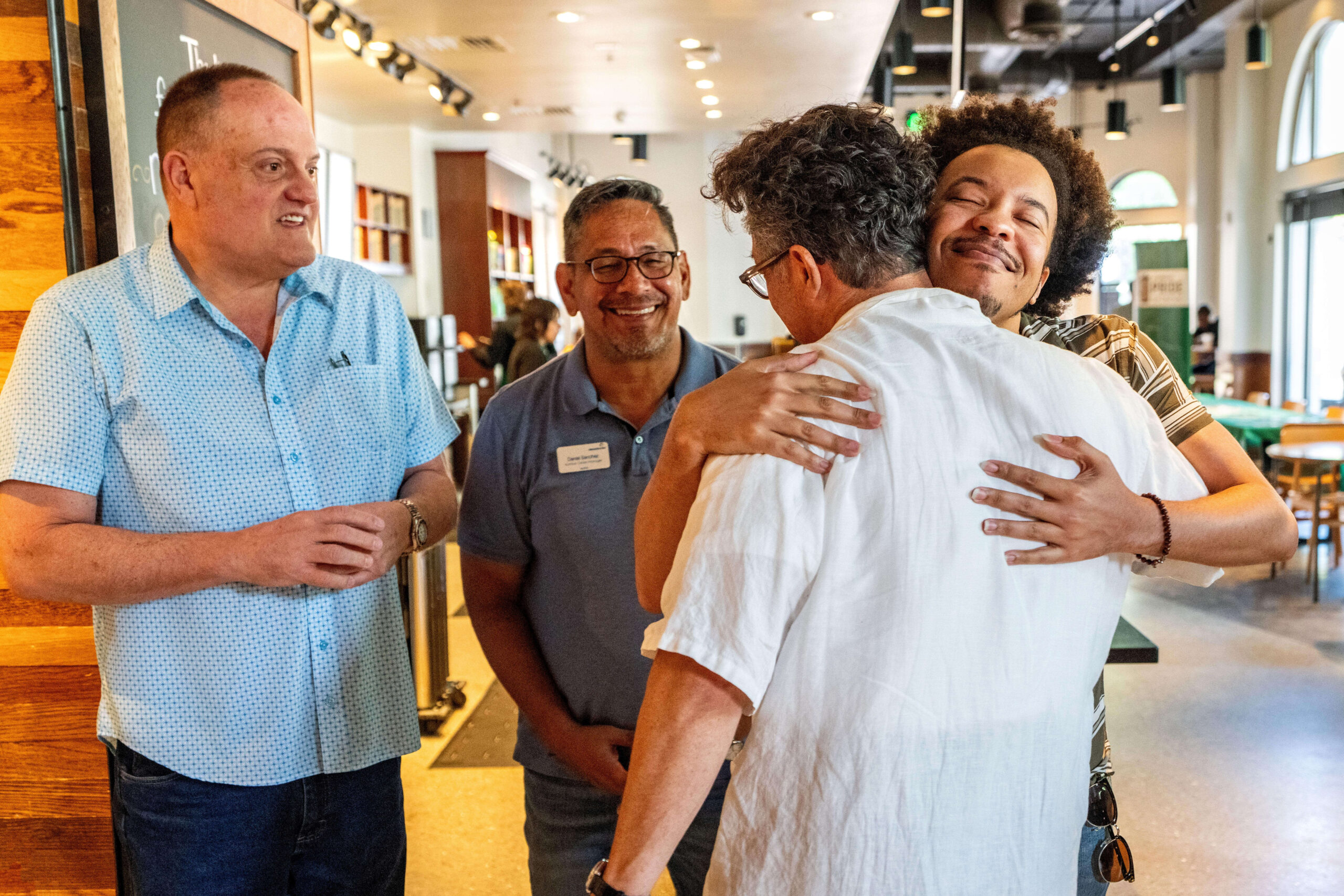 A group of four people is interacting in a warm, friendly setting. Two individuals are hugging, while the others smile and watch. The atmosphere appears joyful and welcoming.