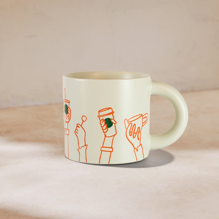 A light green mug features playful orange line drawings of hands holding various drinks, including a coffee cup and a straw.