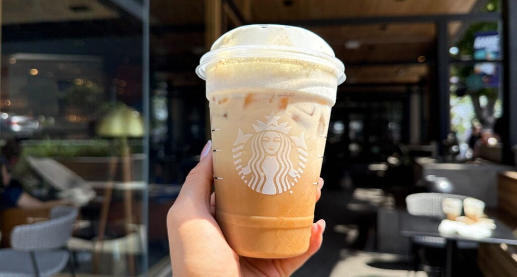 A hand holds a clear Starbucks cup filled with iced coffee, topped with foam. The background features a café setting with outdoor seating.