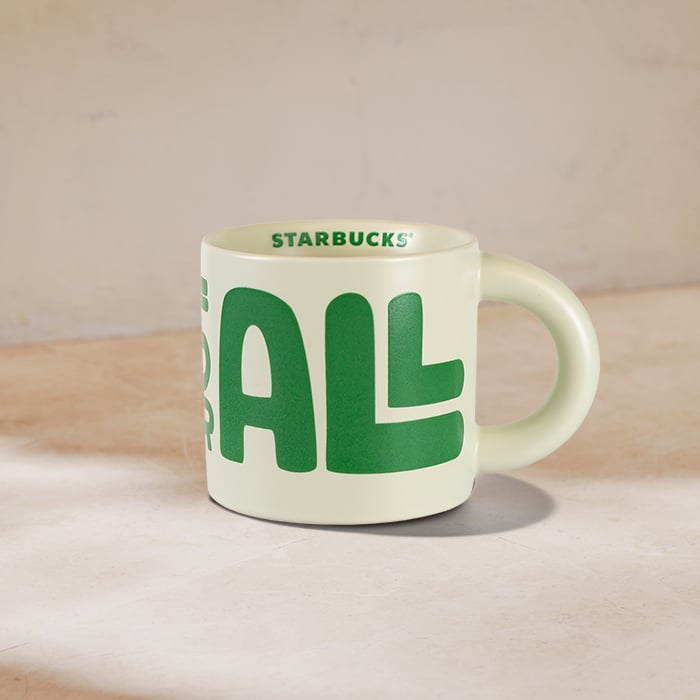 A light green Starbucks mug features bold green lettering that reads "FOR ALL." The mug has a rounded handle and a smooth finish.