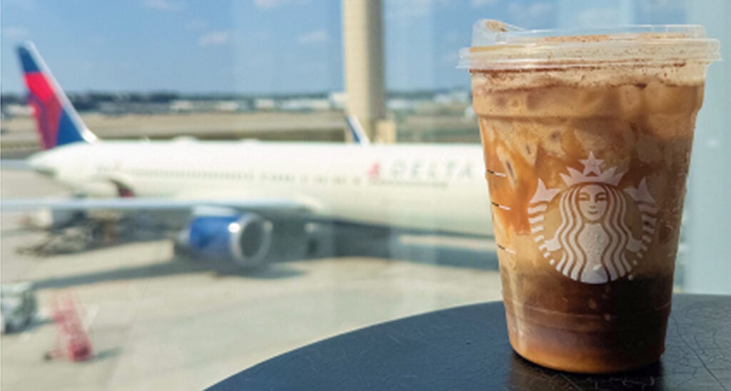A Starbucks iced coffee sits on a table in the foreground, with a Delta Airlines airplane visible in the background at an airport.