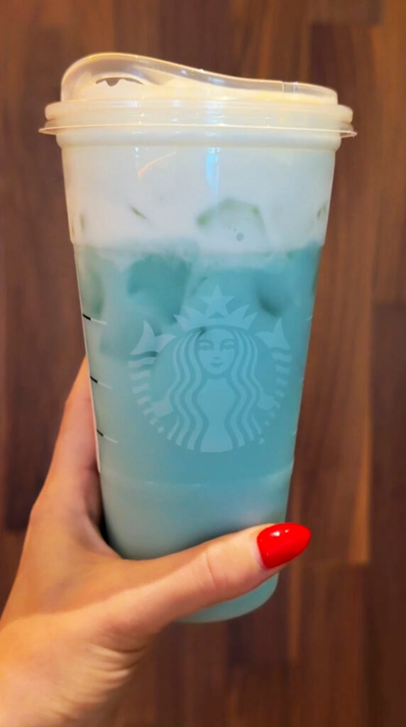 A hand holds a clear cup filled with a blue beverage and topped with white foam. The background features a wooden surface.