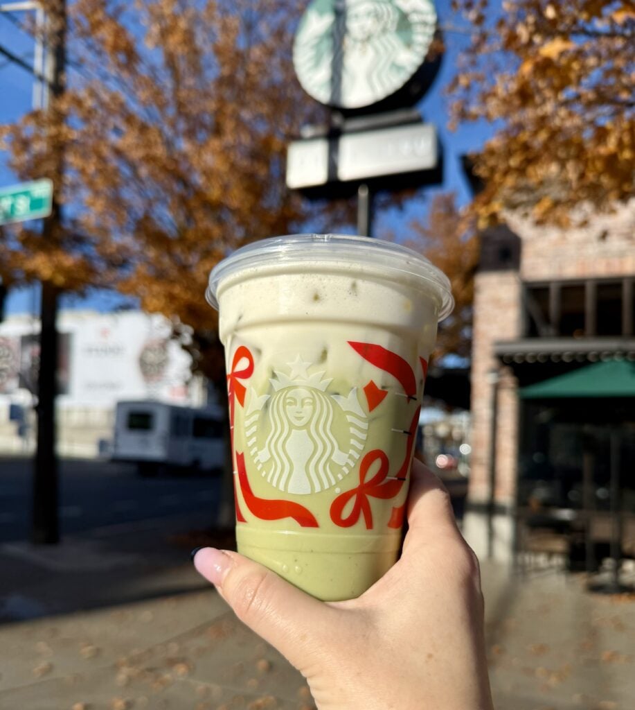 A hand holds a Starbucks cup filled with a green beverage, featuring festive red decorations. In the background, a Starbucks sign and autumn leaves are visible.