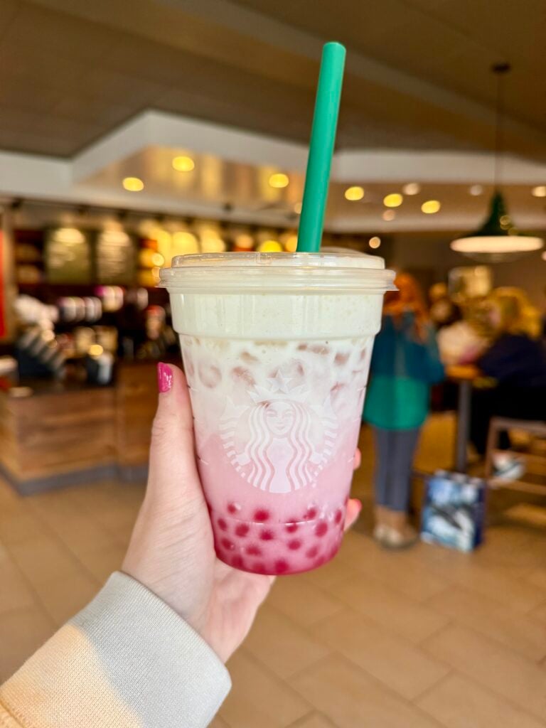 A hand holds a Starbucks drink with a green straw. The beverage features a layered appearance with pink and white colors, and pink tapioca pearls at the bottom. The background shows a cozy café setting.