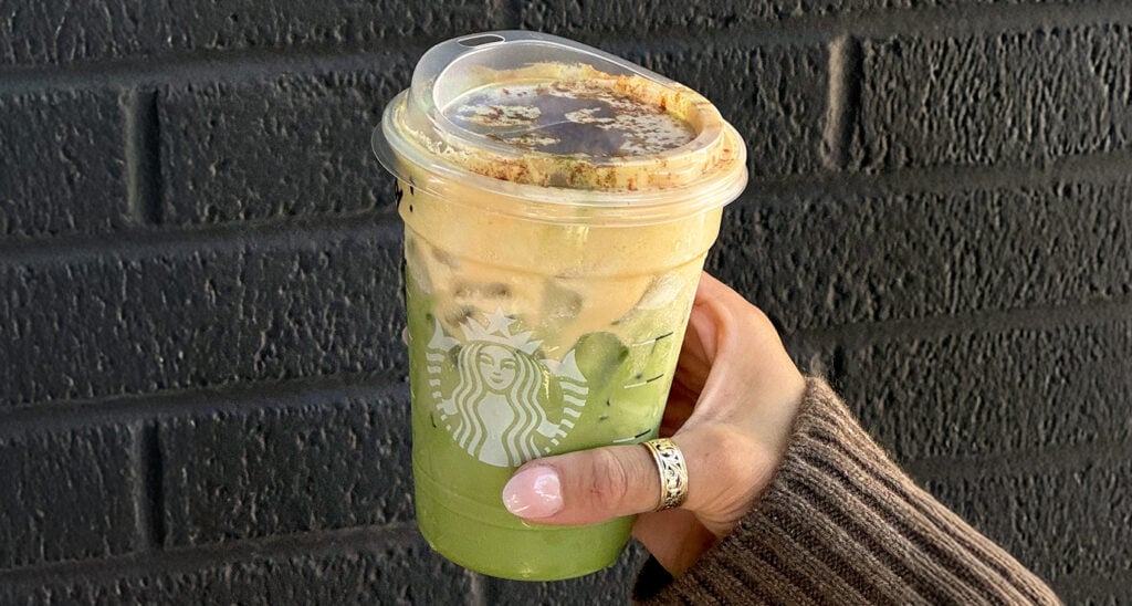 A hand holds a clear cup of layered green and brown beverage, topped with foam and a sprinkle of cinnamon, against a textured black wall.