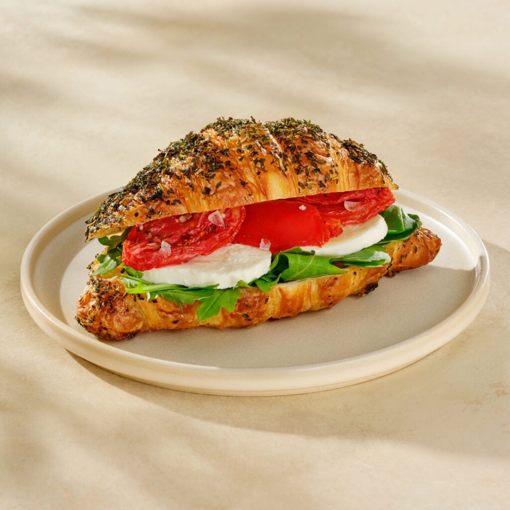 A croissant sandwich filled with fresh mozzarella, arugula, slices of tomato, and topped with herbs, served on a plate.
