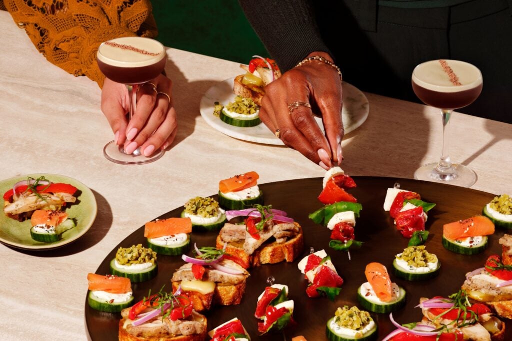 A variety of appetizers are displayed on a dark platter, featuring cucumber slices topped with cream cheese, smoked salmon, and colorful garnishes. In the foreground, a hand reaches for a skewer with assorted toppings, while another hand holds a cocktail. The setting has a vibrant, inviting atmosphere.