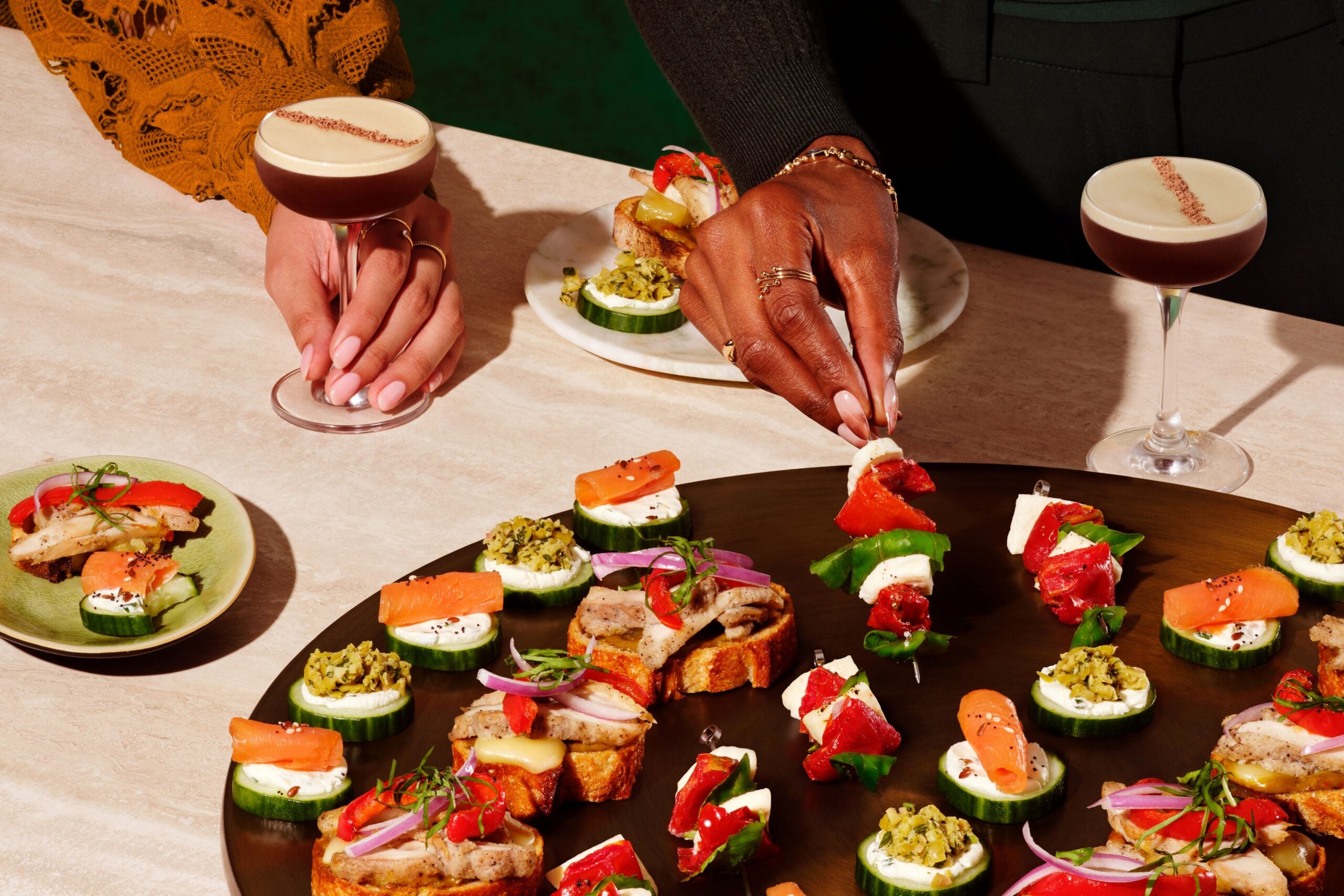 A variety of appetizers are displayed on a dark platter, featuring cucumber slices topped with cream cheese, smoked salmon, and colorful garnishes. In the foreground, a hand reaches for a skewer with assorted toppings, while another hand holds a cocktail. The setting has a vibrant, inviting atmosphere.