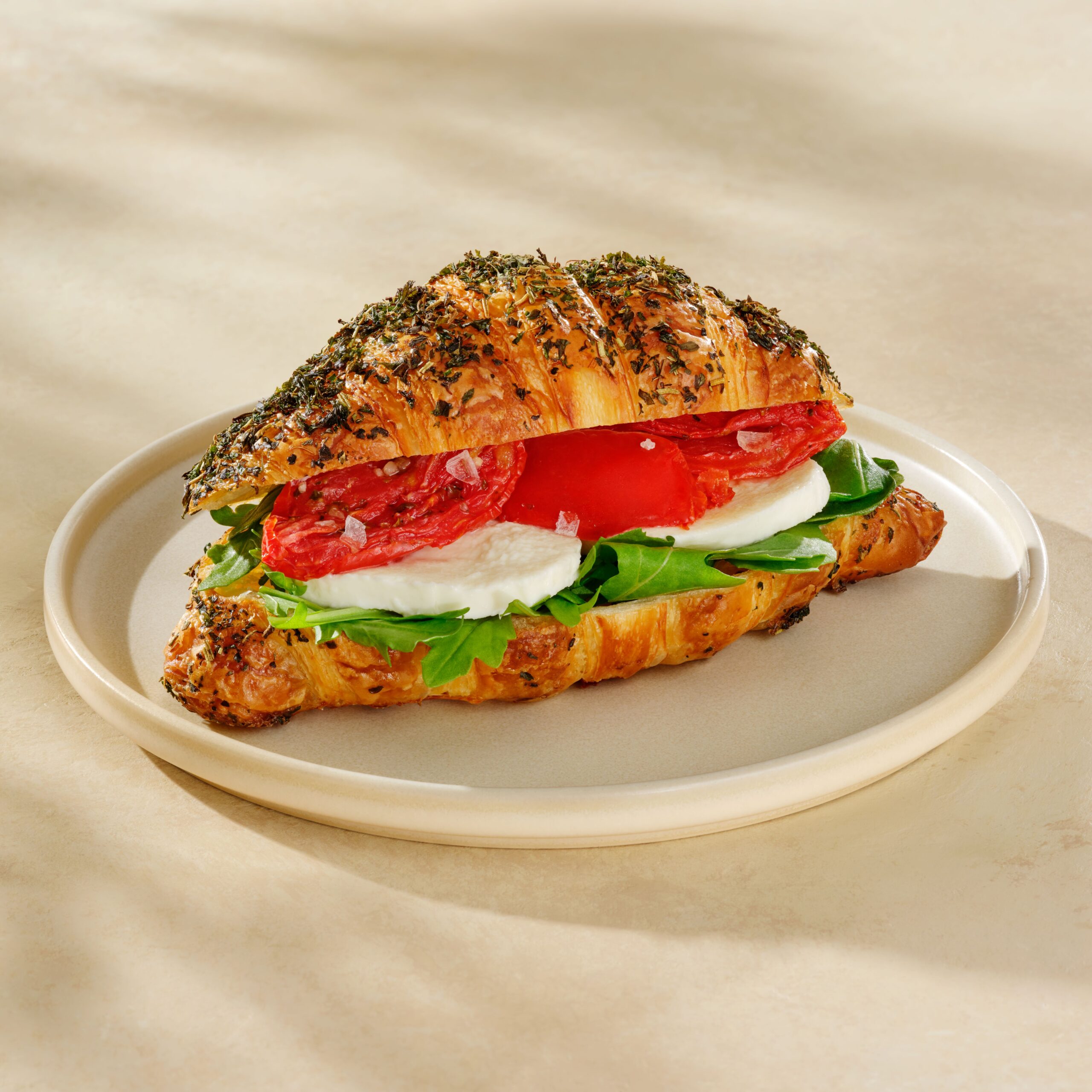 A croissant sandwich filled with fresh mozzarella, arugula, slices of tomato, and topped with herbs, served on a plate.