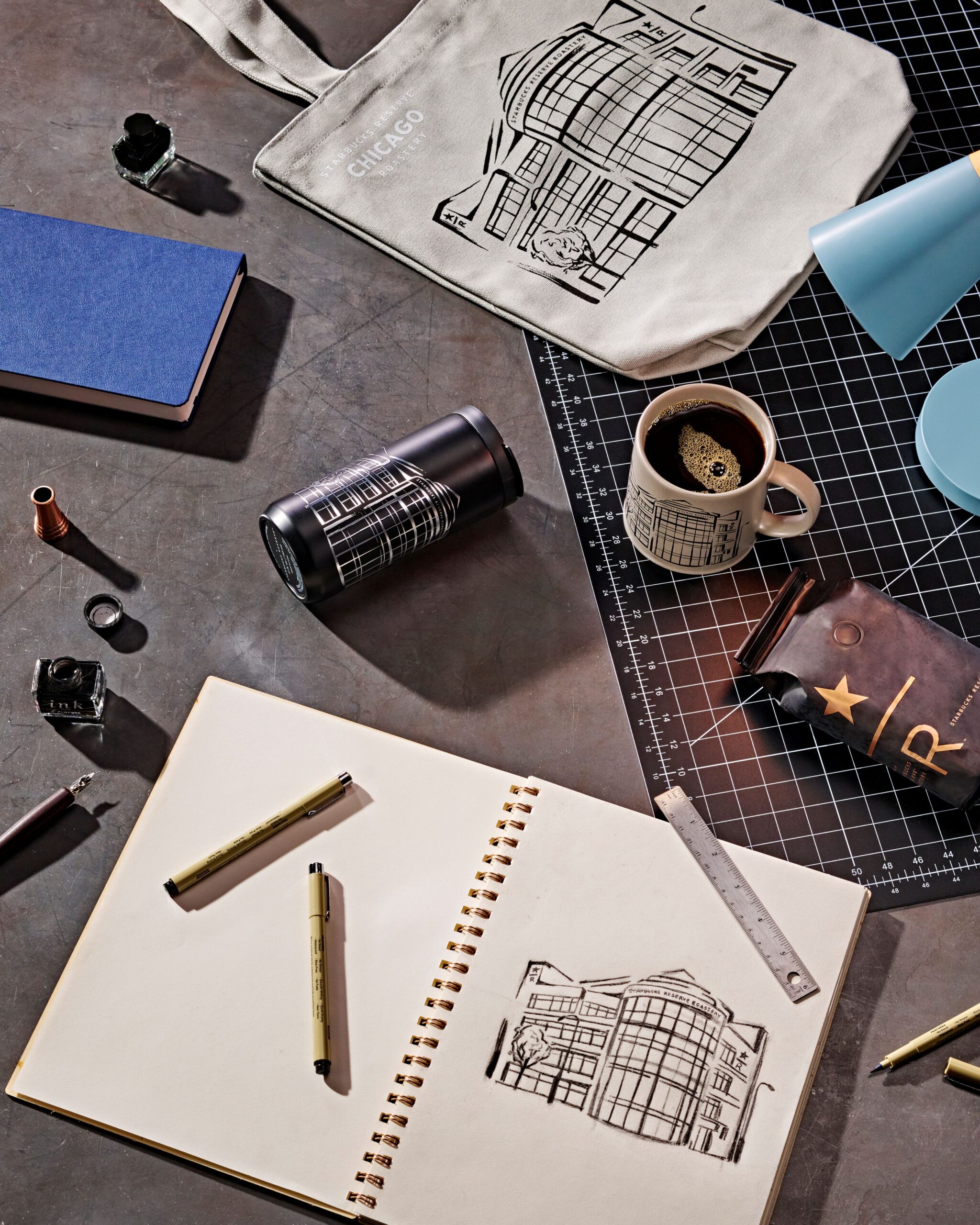 A flat lay of art supplies and a sketchbook featuring a building drawing. Items include a tote bag, pens, a notebook, a coffee mug, and a ruler.