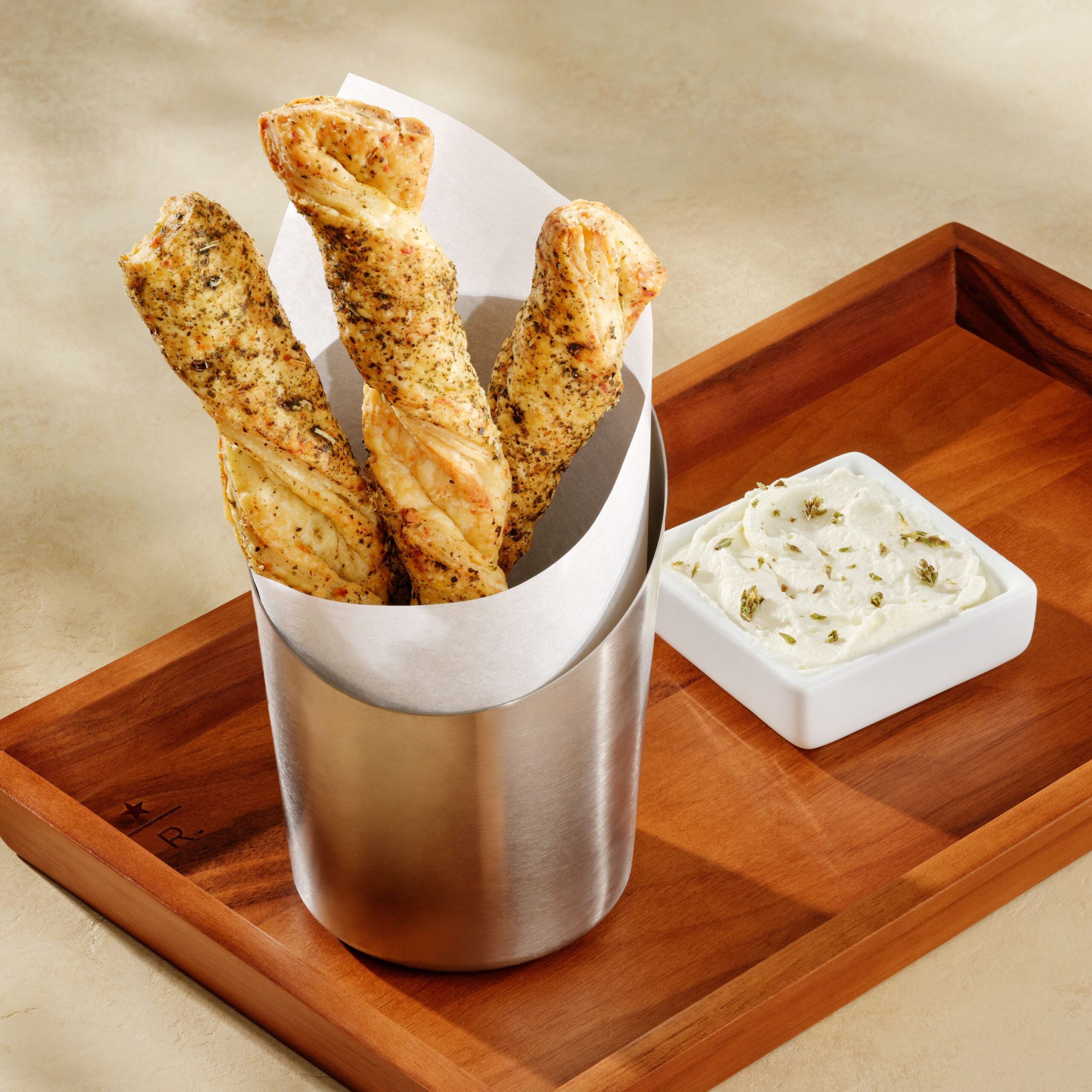 Three twisted breadsticks, seasoned with herbs, are served in a silver container lined with paper. A small dish of creamy dip is beside them on a wooden tray.