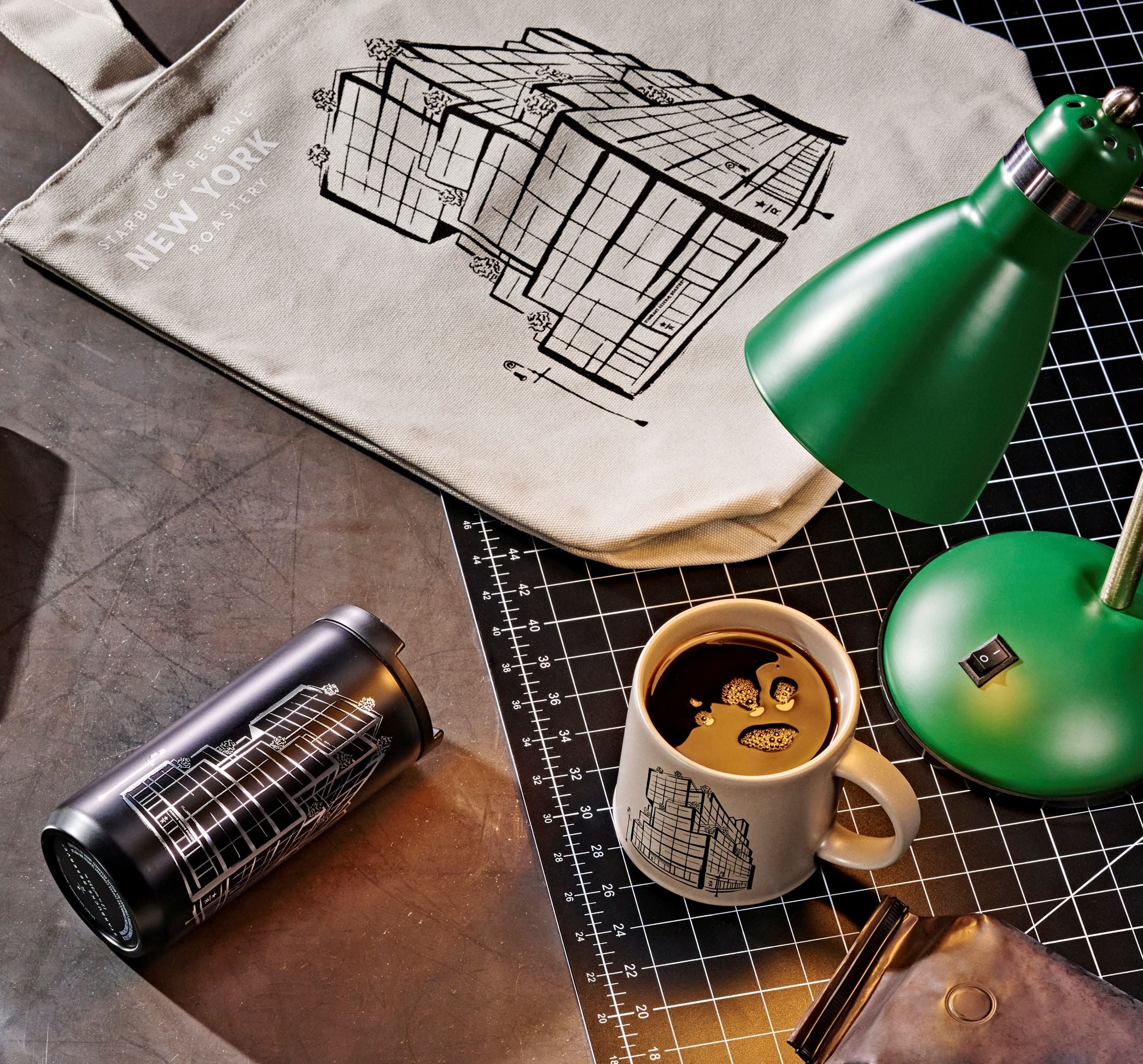 A stylish workspace featuring a beige tote bag with a building illustration, a green desk lamp, a black travel mug, and a coffee cup with a similar design. A brown leather wallet is also visible on a grid-patterned surface.