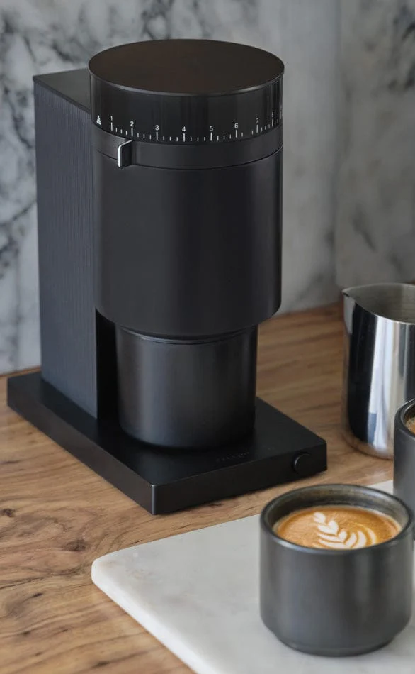 A sleek black coffee maker sits on a wooden surface, with a cup of latte art beside it. The machine features a round top with measurement markings.