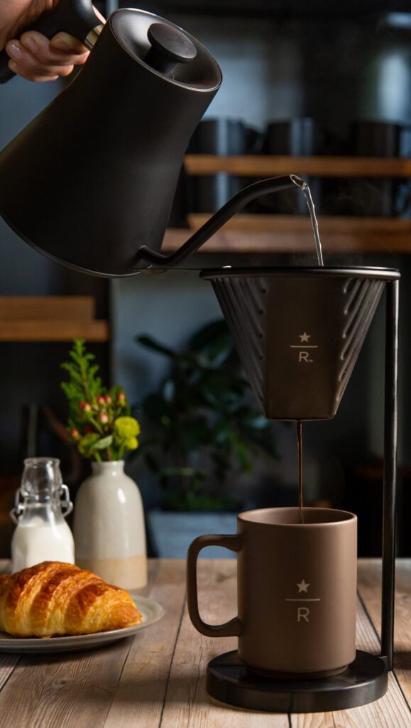 A black kettle pours hot water into a coffee dripper, with brewed coffee flowing into a brown mug. A croissant and milk in a bottle are nearby.