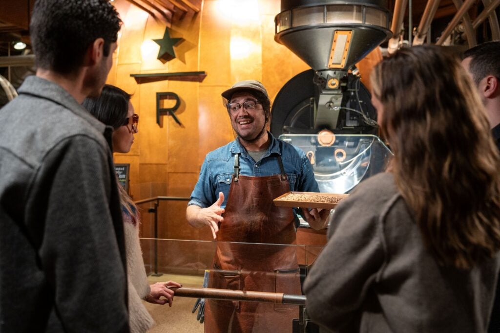 A barista in a denim shirt and brown apron is engaging with three customers, explaining coffee beans while holding a tray. A coffee roaster is visible in the background.